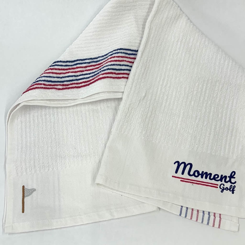 Moment Golf white cotton caddy towel with red and blue stripes, embroidered flag logo, and Scripted branding.